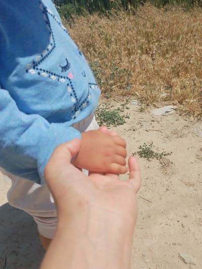 The image shows a close-up of two hands, one adult and one child, clasped together outdoors. The child's hand, which is smaller and has a darker skin tone, is held within the adult's larger, lighter-skinned hand. The child is wearing a blue long-sleeved shirt with a white and dark blue embroidered pattern that resembles a cartoon face with closed eyes and pink cheeks. The background consists of dry, yellowed grass and sandy ground, suggesting a rural or arid environment. There are also some green plants and rocks scattered in the background. The overall lighting indicates a sunny day. There is no indication of the location in the city of Mady, Kyrgyzstan.