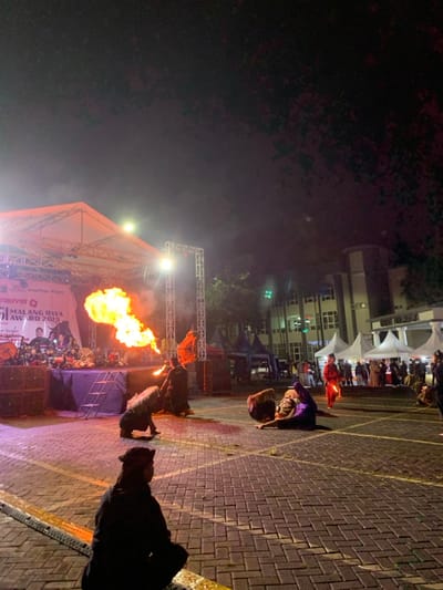 The scene is a nighttime outdoor performance in Kecamatan Batu, Indonesia, at the coordinates provided. A large flame shoots out from a fire-breathing performer standing on the stage, while several other performers wearing elaborate costumes and traditional masks are situated around the stage. The stage is made of metal and wood and is set up in front of a larger building with several windows. It is set up in front of the building. The performer is facing away from the camera, and the flame is directed toward the right side of the image. To the left of the stage, there are spectators seated on the ground. The performer is wearing a black costume with a red sash. The stage is decorated with a banner that reads "MALANG RAYA AWARD 2023". The image appears to have been taken during a nighttime performance.