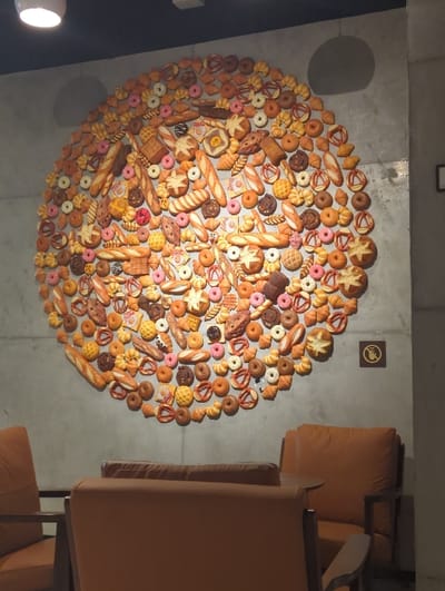 A collection of bread, pastries, and donuts are arranged in a circular pattern on a gray wall. A no-smoking sign is positioned above the arrangement. To the right, orange armchairs are visible, partially obscuring the artwork. The lighting appears dim, highlighting the textures and colors of the baked goods.