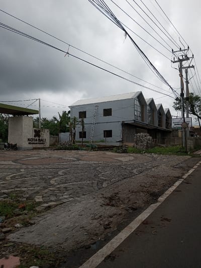 This image depicts an outdoor scene in Vaatharawattai, Sri Lanka, under a heavily overcast, dark grey sky.
The foreground consists of an asphalt road on the right with a visible white edge line. Adjacent to the road on the left, a patterned paved area, composed of interlocking bricks in circular and curvilinear designs, forms an entrance or forecourt. Small patches of weeds and dirt are visible along the road's edge and within the paving cracks.
In the midground, to the left, a light-colored, possibly concrete, entrance structure with a dark green canopy is present. A sign on this structure displays a butterfly logo and the text "KOTA BALI MEKASAR RESORT." A dark-colored motorcycle is parked next to this entrance. The central and right midground features a complex of light grey buildings with distinctive gabled roofs, appearing to be in a state of construction or renovation. The leftmost building section is two stories with four empty rectangular window openings, against which a silver extension ladder leans. Adjacent to it are three further sections, each with a steep gabled roof and large, multi-paned window structures on the upper level. The lower levels of these sections have closed, dark-colored panels or doors. Stacks of bricks or masonry blocks are located on the ground in front of these buildings. To the far right, a white sign on a pole reads "MARKETING OFFICE" with additional, partially obscured text below it. Scattered trees and shrubs are visible behind the entrance structure and between the buildings.
The background is dominated by the dense, dark grey clouds. A network of black electrical or communication wires crisscrosses the upper part of the scene. Multiple utility poles are present, including a substantial pole structure with crossarms and insulators on the far right. A small, red and white object, possibly plastic or fabric, is entangled in one of the overhead wires in the upper left portion of the image. No individuals are visible, and no active interactions are occurring.