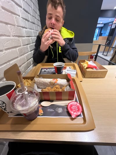 The image depicts a young man sitting at a table in a fast-food restaurant in Jiříkovice, Czechia. He is wearing a black jacket with a yellow hood and is eating a burger with his right hand. The table is covered with a wooden laminate and is cluttered with food items and empty packaging. The man's left hand is resting on the table. In front of him are two trays. One tray is filled with a wrap, fried chicken pieces, french fries, a cup of a purple-colored drink, a cup of coffee, and a small cup of KFC sweet chili sauce. The other tray has a box of KFC fried chicken, a few french fries, and a paper cup. The wall behind him is made of white brick. The restaurant has light brown tables and black metal chairs. The restaurant is brightly lit and a man dressed in black can be seen standing further away in the background. The scene is casual and relaxed, the man appears to be enjoying his meal.