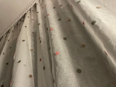 The scene, located in Vlahovo, Bulgaria, captures an upward view of a white, semi-sheer curtain adorned with textured, multicolored polka dots. The curtain hangs in vertical folds, revealing a visible weave pattern in its fabric. The circular dots are distinct and raised, appearing in shades of greenish-grey, pink, gold, and occasional reddish hues, distributed across the fabric surface. Above the curtain, a light-colored ceiling meets a decorative white crown molding or trim piece, which features a repeating, ornate relief pattern. The curtain is suspended from a concealed track system positioned beneath this molding. Ambient lighting casts soft shadows within the curtain's folds. No individuals, actions, or specific interactions are depicted.