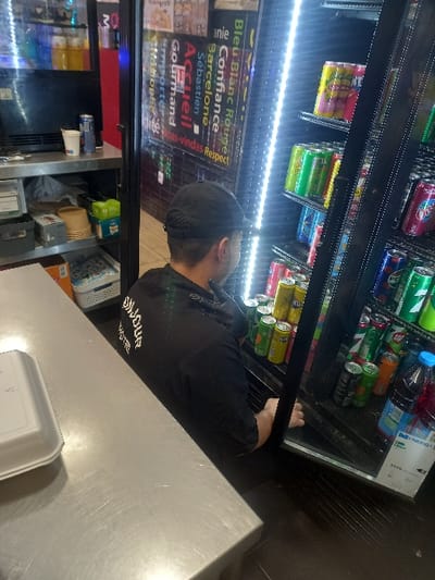 A person wearing a black shirt and a cap is seen standing in front of a refrigerated display case filled with various canned and bottled beverages. The display case also features illuminated signs in French and other languages. In the foreground, a stainless steel countertop is visible, with a white container on it. The environment suggests a retail or convenience store setting.