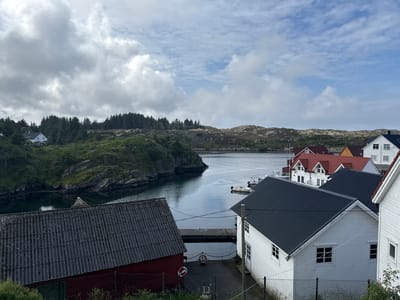 This media file depicts a scenic view of a coastal village in Tjeldstø, Norway. The scene is dominated by traditional Scandinavian architecture, with several white wooden houses featuring dark-pitched roofs. One house in the foreground has a corrugated grey roof and a red lower section. Adjacent to the houses, a calm body of water, likely a fjord or bay, reflects the overcast sky.
The environment is characterized by rocky, green hillsides dotted with coniferous trees in the background. A small pier or jetty extends into the water. A few boats are visible, including a white motorboat moored near the shore. The weather appears to be cloudy with patches of blue sky, suggesting a mild, possibly cool, day. The time of day is likely daytime, given the lighting conditions.
The overall impression is one of tranquility and rural charm. There are no people visible in the frame, and the scene is still and peaceful. The location cues, such as the architectural style and the coastal landscape, strongly suggest Norway. There is no visible text in the image.