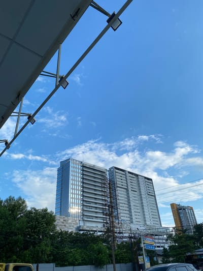 The image captures a bright, daytime scene in Sungai Lakum, Indonesia, with a clear blue sky dotted with white clouds. The foreground is dominated by a large, angled white canopy structure, supported by metal beams and illuminated by industrial lights. Below this canopy, two tall buildings rise into the sky. The building on the left is modern, featuring a glass facade that reflects the sunlight. To its right stands another, older high-rise with a pale, white exterior and numerous balconies. In the middle ground, lush green trees form a dense line across the frame, obscuring the lower parts of the buildings. A yellow vehicle is partially visible at the bottom left corner. The background shows more buildings, including a distinctive yellow and grey tower labeled "GRAND TAN". Electrical wires and utility poles are also present, indicating an urban environment. The overall impression is one of a developed urban area under a clear sky.