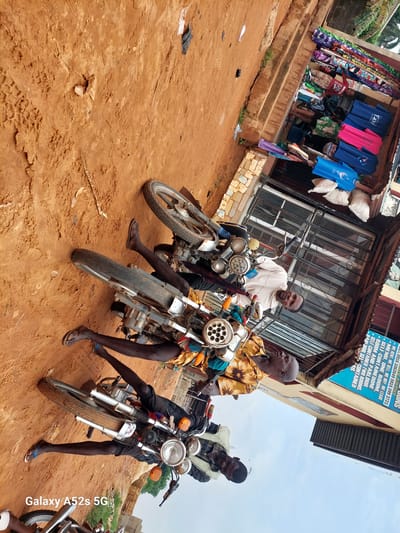 The image, taken in Umudioka, Nigeria, depicts a street scene with several motorcycles and individuals. The ground is covered in reddish-brown dirt, with tire tracks and scattered debris. In the background, a building with a weathered facade houses a shop selling colorful fabrics and t-shirts, some bearing the word "FLOW". Two men are prominently featured. One, wearing a light-colored hoodie and black pants, is seated on a motorcycle, looking towards the camera with a slight smile. Another man, wearing a yellow shirt with a floral pattern, is also on a motorcycle, looking upwards. Both men appear to be of African descent. Other motorcycles are parked nearby, and the overall atmosphere suggests a commercial area. The lighting indicates it is daytime.