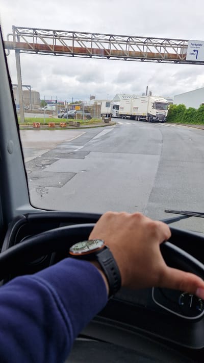 The image is taken from the interior of a vehicle, looking out through the windshield. A person's hands are visible, one on the steering wheel and the other resting on the armrest, wearing a dark blue long-sleeved shirt and a smartwatch. Outside, a road leads towards several parked trucks. A sign visible in the upper right reads "Parkplatz Parking Space 110 M". The environment appears to be an industrial area with buildings and industrial infrastructure in the background. The sky is overcast. The specific location within Essen, Germany, is not identifiable from the provided image.