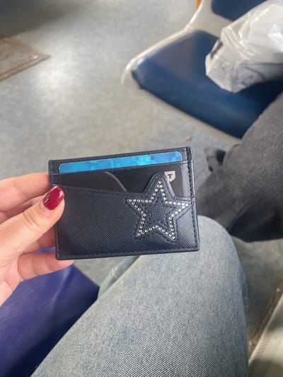 A person is holding a dark blue cardholder adorned with a star made of rhinestones. Two cards are visible inside the cardholder: one with a blue and white pattern, and another black card with a white "T" logo. The background suggests an indoor environment with blue seating and a gray floor. The image does not provide any specific clues to determine the location in Aprelevka, Russia.