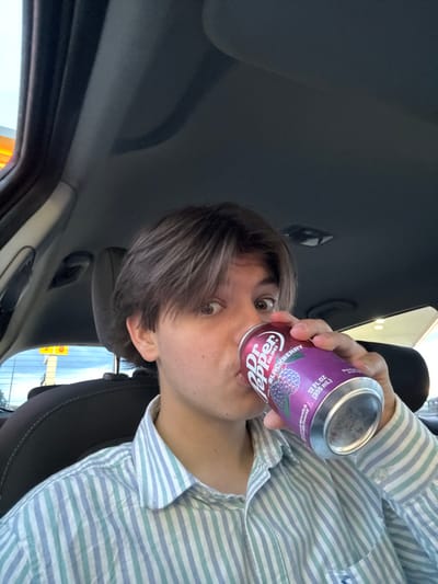The image shows a young adult male, with brown hair, sitting in the driver's seat of a car. He is wearing a striped shirt and is drinking a can of Dr Pepper, blackberry flavored. The can is being held in his right hand, and his left hand is not visible. The car is parked in a gas station in Pocklington, United Kingdom (latitude: 53.921, longitude: -0.796). The interior of the car is visible, and the exterior can be seen through the windows. The windows are clean, and the car is in good condition. The image was taken from a first-person perspective, with the camera held at a slight angle. The lighting is good, and the image is in focus. The scene is typical of a gas station parking lot, and the overall tone is casual and mundane. The person is drinking their beverage in a relaxed manner. The image was likely taken in the afternoon or early evening, as the light is not strong and there are no shadows visible. There is no evidence of other people in the image.
