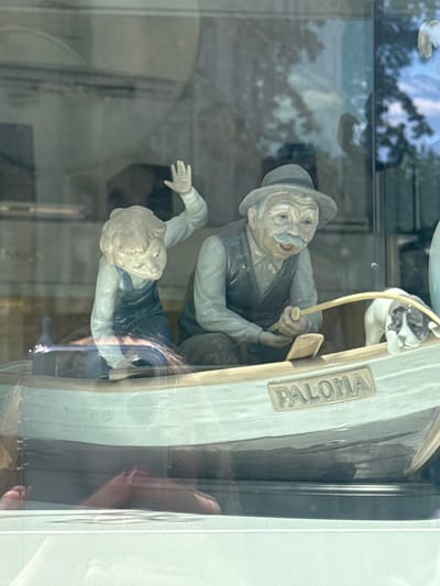 This image captures a ceramic figurine scene through a reflective surface, likely a display case or window. The central subjects are two human figures and a dog, all rendered in a detailed, porcelain-like material. One figure appears to be an older man wearing a hat and vest, with a prominent mustache, holding a fishing rod. Beside him, and seemingly falling or struggling, is a younger figure, possibly a child, wearing a dark vest over a light shirt. A small dog, with black and white markings, is positioned above the man, reaching out with its paws. The figures are depicted in dynamic poses, suggesting an action or narrative.

The scene is set on what appears to be a boat, with the word "PALOMA" clearly visible in raised lettering on its side. The boat's design is light-colored and sleek. Reflections from the surrounding environment, including trees and the sky, are visible on the surface of the figures and the display. The lighting suggests it is daytime, with diffused natural light. The overall mood conveyed is one of slight drama or concern, particularly due to the postures of the figures. The presence of a display case implies this is likely a shop or exhibition setting, possibly in Karlovy Vary, Czechia, as suggested by the location context.