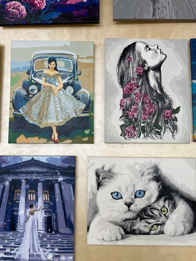 The image displays a collection of five paintings, arranged in a grid-like fashion on a textured, light-colored wall. The paintings appear to be of the paint-by-numbers variety, characterized by their bold outlines and distinct color blocks.
On the top row, a painting on the right depicts two cats, their faces close together. One cat has piercing green eyes, while the other has striking blue eyes. The overall tone is monochromatic with the exception of the eye colors, suggesting a dramatic or striking composition. To the left of the cats, only a sliver of another painting is visible, featuring abstract grey and white shapes.
The bottom row features three paintings. The left-most painting in this row is a close-up of dark purple and blue flowers with touches of pink. The painting to its right showcases a woman in a retro, polka-dot dress lying down in the driver's seat of a vintage car. She wears red shoes. The background is abstract, with muted earth tones and dark green. The final painting in the collection, positioned to the right, shows a nighttime cityscape with a person in a flowing white garment seemingly leaping or dancing between tall, dark buildings. The scene is rendered in shades of purple and blue, with hints of light reflecting from the windows of the buildings.
The paintings are hung indoors, likely in a home or a gallery setting, as indicated by the wall texture and the arrangement. No text is visible on any of the paintings. The overall impression is one of diverse artistic expression, from stylized animal portraits to vintage scenes and urban landscapes.