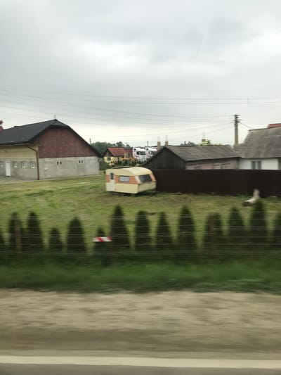 This image presents a roadside view in Milișăuți, Romania, captured on an overcast day from a moving perspective, evidenced by motion blur in the foreground. The sky is uniformly light grey and cloudy, traversed by multiple horizontal power lines in the upper portion.

In the blurred foreground, a light brown/tan earthen or gravel shoulder transitions to a dark grey asphalt road surface. The middle ground consists of a vibrant green grassy field. A row of dark green, conical evergreen shrubs forms a hedge, positioned in front of a dark brown privacy fence that stretches horizontally across the scene. A multi-strand wire fence is visible just in front of the hedge, and a small red and white striped marker is affixed near its base, slightly left of center.

Parked within the grassy field, slightly left of center, is a vintage-style travel trailer or caravan, predominantly beige or light yellow with an orange stripe or panel. Its door and a window are visible on its side.

In the background, behind the grassy field and fence line, various structures are present. On the left, a prominent two-story building features a dark grey gabled roof, reddish-brown upper walls, and a lighter tan/concrete ground floor with multiple rectangular windows and a white door. To the right, obscured partially by the brown fence and trees, are several smaller rural houses with dark grey and reddish roofs. Further back, multiple white trucks, potentially semi-trucks, and a yellow construction vehicle are visible amidst trees. A tall wooden utility pole, equipped with crossarms and wires, stands on the far right. An indistinct, light-colored, blurry object lies in the grass behind the hedge on the right. No discernible human or animal figures are clearly present or engaged in any activity.
