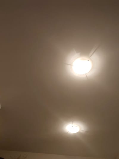 The image shows a ceiling with two illuminated light fixtures. The ceiling is a neutral, light brown color. The upper light fixture is larger and brighter, with a circular diffuser and thin metal supports extending outwards. The lower light fixture is smaller and slightly dimmer, also circular in design. The lighting casts soft shadows on the ceiling. The lower portion of the image shows a hint of a wall with some discoloration or texture. The scene is likely an interior space in a building. There are no people or discernible actions taking place. The specific location in Ravda, Bulgaria, cannot be determined from the visual information alone.