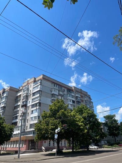 The image captures an urban scene on a sunny day in Zhytomyr, Ukraine. The primary subject is a multi-story apartment building, its grey brick facade and numerous balconies dominating the frame. Lush green trees with dense foliage are strategically placed, partially obscuring the lower floors of the building and lining the adjacent asphalt road. A white car is visible parked on the side of the road, near a sidewalk. Overhead, a network of black utility wires crisscrosses the bright blue sky, punctuated by scattered white clouds. The presence of a traffic light and a pedestrian crossing sign indicates an intersection or a busy street. The perspective is from a low angle, looking upwards, which emphasizes the height of the building and the expanse of the sky. The overall impression is that of a typical residential area in a city, bathed in the clear light of day. No people are directly visible in the image.