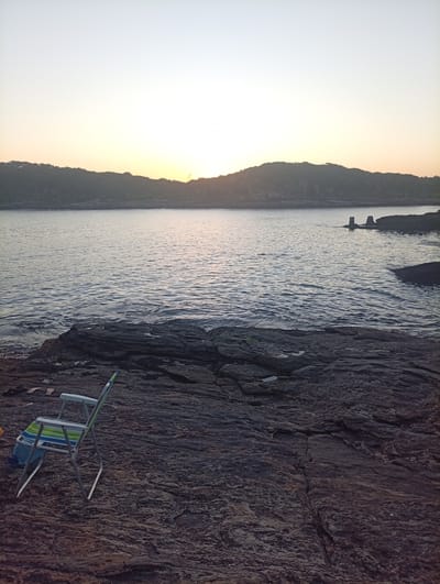 The image captures a serene coastal scene in Armação dos Búzios, Brazil. The foreground features a folding chair resting on dark, rocky terrain that slopes towards the water's edge. The sea stretches towards a distant coastline marked by undulating, tree-covered hills. The sky is a gradient of pale yellows and whites, indicating either sunrise or sunset. The water's surface is gently rippled, reflecting the soft light from the sky. In the middle distance, partially submerged stone structures are visible in the water.