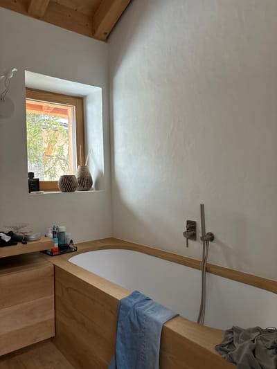 The image shows a bathroom with a white bathtub and a wood surround, located in a house in Celerina/Schlarigna, Switzerland (latitude: 46.514, longitude: 9.859). The bathtub is empty and has a silver showerhead and handle. A blue towel is draped over the side of the tub. Another towel, gray in color, is laying on the floor, next to the tub. There is a wooden shelf above the tub and another shelf to the left, both holding various items. A window is in the wall to the left of the tub and a white bird sculpture is hanging from the ceiling above it. Light from the window is shining into the bathroom. The bathroom is otherwise empty. The walls are white and the floor is wooden.