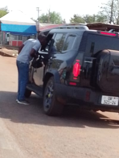 A man stands beside a dark-colored SUV with a rear-mounted spare tire. The vehicle's license plate reads "UW 521-24". The man is dressed in a light blue shirt and blue jeans. He appears to be looking into the vehicle. The location is Dungu, Ghana. A tent with a blue and white awning is visible in the background.