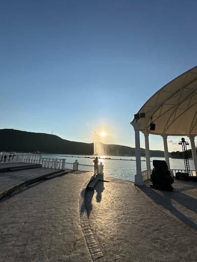 The image depicts a waterfront promenade at what appears to be sunset. The sun is low on the horizon, casting a warm glow and long shadows across the textured pavement. In the foreground, the promenade is lined with decorative white railings and columns, suggesting a developed tourist area. The sea stretches out to a dark, mountainous coastline in the background, partially silhouetted against the bright sky.
A large, white canopy structure with metal supports is visible on the left, providing shade. The water is calm, reflecting the sunlight, and a faint mist or spray rises from the surface, possibly from a fountain or sea spray. A few people are visible, mostly as silhouettes, standing and walking along the promenade, enjoying the view and the evening light.
On the far right side of the image, part of a stage or performance area can be seen, with lights and equipment, hinting at potential events or entertainment. The overall atmosphere is serene and picturesque, with the warm light of sunset creating a tranquil mood. The location is Abrau-Dyurso, Russia, known for its wineries and scenic coastline. The clear sky indicates fair weather.