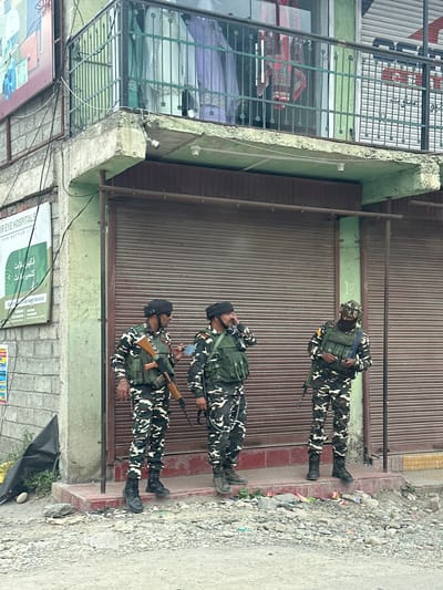 Three individuals in camouflage uniforms, equipped with rifles and tactical vests, stand outside a building with shuttered storefronts. The building appears to be a commercial establishment, possibly a shop or clinic, as indicated by a sign that reads "EYE HOSPITALS FOR BETTER LIFE" in English and Urdu. The individuals are positioned on a raised concrete platform in front of the shutters, which are a metallic brown color and appear to be pulled down. One individual on the left is holding a rifle and looking to their right. The central individual is speaking on a phone and holding a rifle. The individual on the right is standing with their rifle. The ground in front of the building is covered in gravel and debris. The overall environment suggests an urban setting, possibly a street in Cherwan, India, with the presence of security personnel indicating a potentially sensitive or high-alert situation. The building facade exhibits signs of wear and tear, with peeling paint and exposed concrete. Electrical wires are visible running along the exterior of the building.