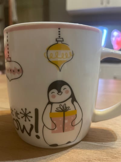 A close-up, eye-level shot shows a white ceramic mug with a pink rim adorned with a festive penguin design. The penguin is holding a pink gift box with a yellow ribbon. Above the penguin are decorative ornaments. Text that reads "Let it snow!" is visible at the bottom of the mug. The mug sits on a wooden surface. The background is blurred, showing what appears to be an indoor setting with cabinets and what might be a television screen with colorful lights. There is no indication of the city of Baku, Azerbaijan in this image.