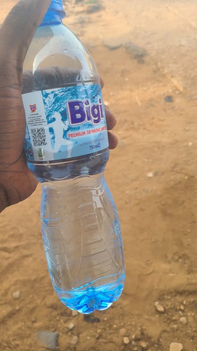 A dark-skinned person holds a bottle of Bigi Premium Drinking Water in their hand. The bottle is mostly clear, with a blue label that features the brand name in large white letters. The label also indicates the volume of the water as 750ml. The bottle is capped with a blue lid.
The background is a dirt or sandy surface with sparse vegetation. The picture was taken in the Mandate Estate area of Ilorin, Nigeria.