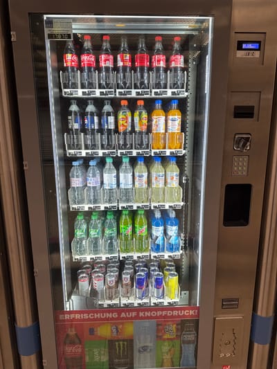 The image shows a vending machine stocked with various beverages. The machine has multiple shelves displaying bottles and cans of soda, energy drinks, and juices. Some visible brands include Coca-Cola, Lipton, Mountain Dew, Pepsi, Fanta, Sprite, and Red Bull. Prices are displayed below some of the items, ranging from 1.50€ to 2.20€. The machine features a digital display showing "EUR 0.00 BETRIEBSBEREIT" (EUR 0.00 READY FOR OPERATION). A keypad and coin slot are visible on the right side of the machine. A sign at the top of the machine has text that includes "GEILE" and a phone number, indicating the machine is serviced by GEILE Getränkeautomaten, located in Gartenkamp 110-112, 49492 Westerkappeln. The bottom of the machine has a large poster with the text "ERFRISCHUNG AUF KNOPFDRUCK!" (refreshment at the push of a button!) and images of various drinks. The overall environment appears to be an indoor public space, possibly a hallway or waiting area, given the metallic walls and floor. No people are present in the image.