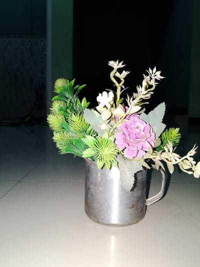 A close-up, eye-level, indoor shot features a bouquet of artificial flowers in a metallic mug. The mug, made of a tarnished silver material with a visible handle, sits on a light-colored, tiled surface. The bouquet consists of various green, spiky foliage, a prominent pink rose, and delicate white flowers, some of which are elongated and wispy. The background is dark and out of focus, with hints of vertical structures and patterned textures. The lighting suggests a dimly lit environment. This scene is located in Sendangrejo, Indonesia.