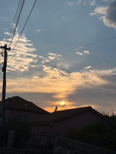 The image depicts a sky filled with clouds at sunset, casting a warm golden glow. A power line cuts diagonally across the frame, with a power pole visible at the bottom. To the right, the roof and side of a building are silhouetted against the bright sky, with some greenery and a fence also visible. The setting appears to be residential, and the time of day is clearly sunset. The clouds are wispy and scattered, some reflecting the sunlight and appearing bright white, while others are tinged with orange and yellow. The overall mood is serene and peaceful, characteristic of a sunset scene. There is no visible text in the image.