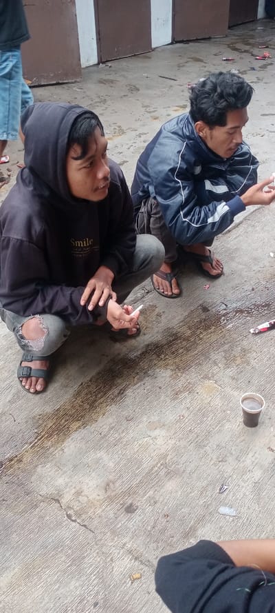The image depicts an outdoor scene in the city of Cilampeni, Indonesia. Two young adult males are observed in a squatting position on a light gray concrete surface. The male on the left wears a black hooded sweatshirt with "Smile" text, light-grey ripped jeans, and dark sandals, holding a white cigarette in his right hand while looking towards the right. The male on the right wears a dark blue and black jacket with white piping, dark pants, and dark flip-flops, looking downwards.

The concrete ground exhibits a textured finish, multiple cracks, and various dark and light stains, including a prominent dark, elongated streak. Litter consisting of small wrappers and debris is scattered across the ground. A clear plastic cup containing a dark brown liquid is present on the concrete in the lower right portion of the frame. The background features a solid dark brown wall or fence interspersed with vertical white sections. The lower legs and feet of a third individual, dressed in light blue jeans and red footwear, are partially visible on the far left. A dark sleeve is partially visible at the bottom right. The scene is under natural illumination.