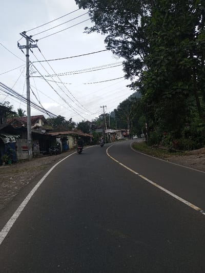 This image depicts a two-lane asphalt road curving to the right in Khajuripada, India, under an overcast sky. The road features a solid white line on its left edge and a broken yellow line with white reflectors on its right, indicating a division or lane boundary. Two motorcyclists, wearing dark clothing and helmets, are visible traveling away from the viewer on the road.
On the left side of the road, a dirt shoulder precedes a line of single-story structures, which appear to be residential or small commercial buildings with varying roof types, including corrugated metal and red tiles. Several utility poles with complex networks of overhead electrical and communication wires stand along this side. Multiple motorcycles are parked in front of these structures.
The right side of the road is bordered by dense green vegetation, including large trees with heavy foliage. Beyond these trees, a forested hilly or mountainous terrain is visible. Another utility pole stands further down the road on the right. A white vehicle is partially visible in the distance, following the curve of the road. No pedestrians are immediately visible.