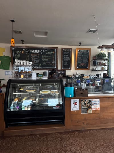 The image shows the interior of a cafe, possibly in South Padre Island, United States, based on the "Best Coffeehouse" sign. The cafe has a menu board on the wall listing drinks, crepes, sandwiches, and ice cream. A refrigerated display case contains pastries and desserts. A person is working behind the counter. Posters and signs are hung on the wooden paneling above the display case. There is a rack of cups and plants above the counter. The cafe appears to have a rustic, casual atmosphere.