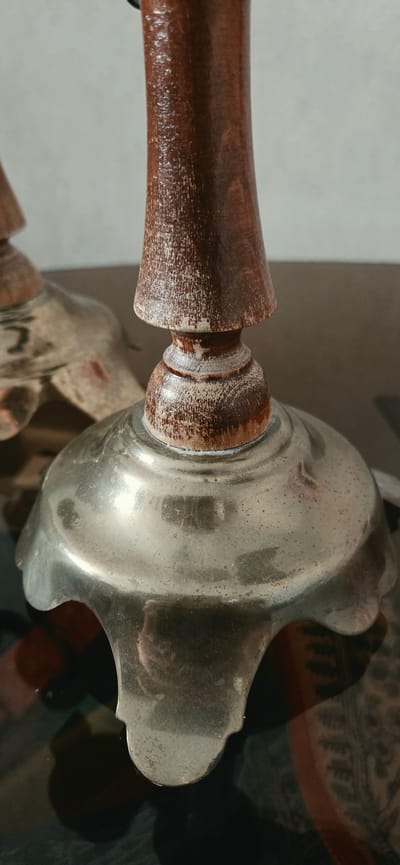 The image shows a close-up, low-angle shot of the base of an antique wooden and metal object, possibly a candlestick or a finial, resting on a dark, reflective surface. The object features a dark brown, varnished wooden top that tapers downwards, with visible wood grain and some white, worn areas where the finish has chipped. This wooden section transitions to a metal base with a scalloped edge and a slightly convex shape. The metal appears to be aged and somewhat tarnished, with visible smudges and light reflections. The background is out of focus, showing a light-colored wall on the left and a darker, patterned surface on the right. There are no people or actions depicted in the image. The specific location in Lipetsk, Russia, cannot be determined from the visual information provided.