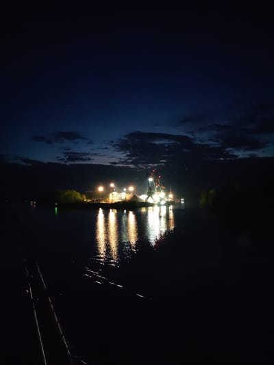 The photograph depicts a nighttime scene in Александровское, Russia, featuring a body of water with a brightly lit industrial area on the opposite shore. Several cranes with red booms are visible within the lighted area. The lights from the industrial area reflect in the water, creating shimmering streaks of light. The sky is dark blue with some clouds. Fishing rods are visible in the lower left of the image. The foreground is dark, with little to no detail visible.