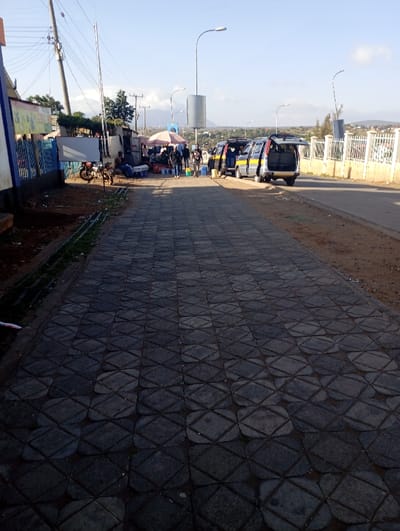 This image captures a bright, sunny outdoor scene in what appears to be a bustling town environment, likely Namanga, Kenya. In the foreground, a wide, dark grey paved walkway extends from the bottom of the frame towards the mid-ground. The pavement features an intricate, interlocking pattern of slightly rounded, diamond-like or octagonal shapes.
To the left of the walkway, there are several simple structures or market stalls, some with blue fencing. A motorcycle is parked near these stalls. A few individuals are visible around this area; one person is sitting on the ground. Further along, a large white umbrella or canopy shades a market stall where several people are gathered, possibly engaged in buying or selling.
On the right side of the frame, a dirt shoulder separates the walkway from a paved road. Several grey vans or minibuses, typical of East African "matatus," are parked along the road. Most of these vehicles have their rear or side doors open, suggesting activity like loading passengers or goods. Near the vehicles and stalls, several blue and white jerry cans or buckets are present on the ground.
In the background, a white metal fence lines the roadside, and a few streetlights with large, cylindrical fixtures are visible. Power poles and wires crisscross the sky. Distant mountains or hills can be seen under a clear, light blue sky. The bright sunlight casts distinct shadows, indicating either morning or late afternoon. On a building or stall to the far left, a colorful sign with indecipherable text is present. The overall scene depicts everyday commercial and transportation activity in a semi-urban setting.