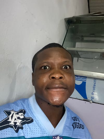 A close-up portrait of a Black man in a light blue jersey with white accents and a "KC" logo. The jersey also displays "GOD'S KINGDOM". He is centered and looking directly at the camera with a slight smile. The background includes a stainless steel object, possibly a refrigerator, and a white wall. The image has been rotated 90 degrees counterclockwise.