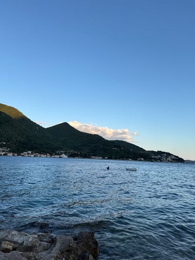 The image depicts a serene coastal scene in Lepetane, Montenegro, under a clear blue sky. In the foreground, a rocky shore meets the calm, blue water of the bay, with gentle ripples disturbing the surface. The water extends to the horizon, where a line of lush, green hills forms the backdrop. Along the foothills of these hills, a picturesque town with white-washed buildings is nestled, suggesting a Mediterranean settlement. A few boats are visible on the water, including a small white dinghy and a dark object that appears to be a person swimming or wading. In the mid-ground, a larger white ferry or boat is docked near the shore, and further along the coast, more buildings are scattered across the landscape. The sun casts a gentle light, illuminating the scene and creating soft shadows on the hills.