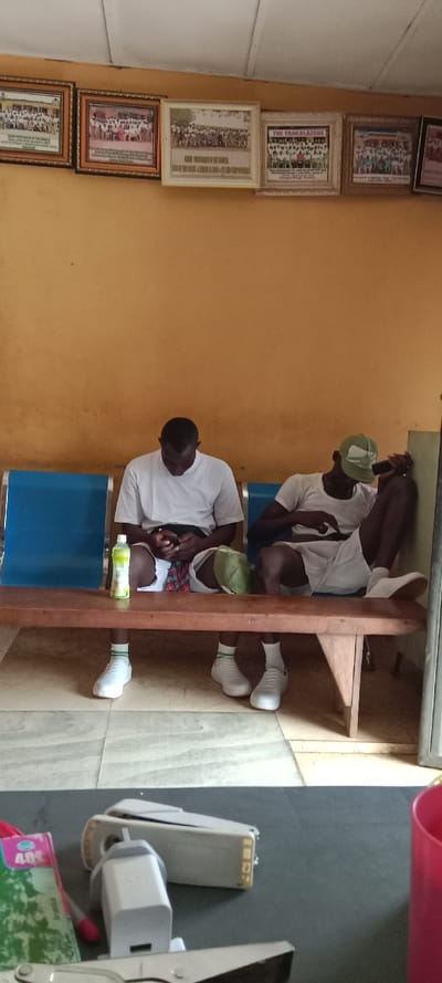 Two men sit on a blue and brown bench, both wearing white shirts, shorts, socks, and shoes. The man on the left looks at his phone, with a bottle of green liquid and a hat on the bench beside him. The man on the right leans against the wall, holding what appears to be a microphone. Above them are framed pictures on a yellow wall. In the foreground, on a dark surface, are office supplies including a stapler, scissors, and a pink cup.