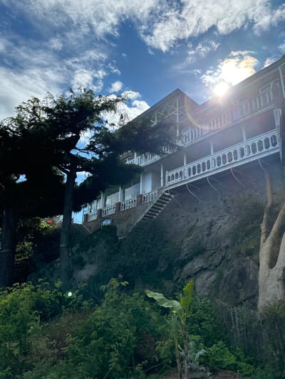 The image shows a white building with multiple balconies built into the side of a rocky cliff.  The building appears to be situated in the  “Le Corsaire” complex located at 48.632, -2.051 in Dinard, France.  There is a staircase leading up to the building, and a large tree in the foreground. The sun is shining brightly in the sky. The building appears to be constructed of white painted wood and has a red brick foundation.  The cliff is covered in vegetation and there are trees in the foreground.