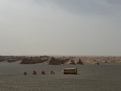 This image depicts a vast, desolate desert landscape in Dunhuang City, China, characterized by numerous striking geological formations known as yardangs. These wind-eroded rock structures, varying in size and shape, are scattered across the mid-ground and extend into the hazy distance, resembling ancient, weathered ruins or a "ghost city." The ground is a flat, sandy, light greyish-brown terrain.

In the foreground, a simple rope fence, strung between dark posts, creates a visual boundary. Inside this fenced area, four prominent red Chinese characters are arranged on the ground. From left to right, they read: 西 (Xī), 海 (Hǎi), 舰 (Jiàn), 队 (Duì). Together, these characters form "西海舰队" (Xī Hǎi Jiànduì), which translates to "West Sea Fleet." To the right of the characters, a rectangular stone block or platform rests next to a wooden fence post, which is part of the rope barrier.

The sky is uniformly pale grey, suggesting heavy overcast conditions or a significant amount of atmospheric haze or dust, common in desert regions. The diffused light eliminates harsh shadows, making the precise time of day difficult to determine, though it is daytime. The overall scene conveys a sense of immense scale, stark beauty, and the profound effects of natural erosion over time. There are no people visible, and no specific activities or events are taking place.