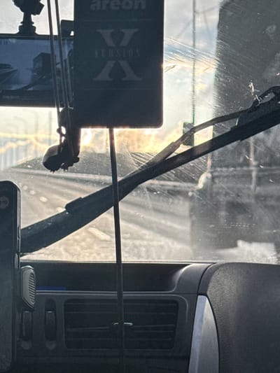 The image displays a view from inside a car, looking out through the windshield, with the dashboard and car interior visible in the lower portion of the frame. The perspective is from the driver's or passenger's seat, looking forward.
In the foreground, a black car dashboard occupies the bottom of the image, featuring air vents and part of a console. A smartphone, possibly resting on the dashboard or in a holder, is partially visible in the bottom right corner, showing a faint "110" on its screen. A car air freshener, black in color and prominently featuring the text "Areon X VERSION" and a large "X" logo, hangs down from what appears to be the rearview mirror area, positioned towards the left side. Wires are visible around this area, suggesting a mounted device such as a dashcam, which is also partially seen. A windshield wiper blade is visible across the upper part of the windshield.
The windshield itself shows significant scratches, streaks, and possibly dirt, which are accentuated by bright sunlight filtering through. This glare creates bright spots and highlights imperfections on the glass. Through the windshield, a blurry outdoor scene is visible, suggesting a road or street with indistinct structures or buildings in the background. The bright sunlight indicates it is daytime, possibly morning or late afternoon given the angle of the glare.
The activity depicted is likely driving or being parked in a car. The overall impression is an ordinary moment from a car journey. The provided location context is Aleksandrovskoe, Russia.