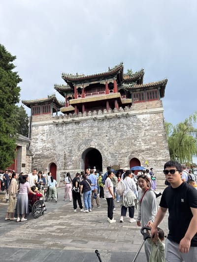 The image depicts a crowded outdoor scene in front of a prominent, ancient-style gate structure, likely a main entrance within the Summer Palace (Yiheyuan) in Haidian District, Beijing, China. The sky above is overcast and bright grey.
The central subject is a large gate building constructed of weathered grey stone blocks, forming a fortress-like base. A large, dark arched entryway is centrally located at the base, with visible stone voussoirs. To the left of the main arch, a smaller, red arched doorway is recessed into the stone wall. Above the stone base, the building features three distinct traditional Chinese pavilion structures with multi-tiered roofs. The roofs are adorned with green glazed tiles, intricate decorative ridge tiles, and red support beams and window frames. The two smaller pavilions flank a taller, central pavilion, all exhibiting ornate architectural details characteristic of imperial Chinese design.
In the foreground and midground, approximately 30-40 individuals, primarily of East Asian descent, are visible. They are casually dressed in modern attire, including t-shirts, blouses, trousers, and skirts. A man wearing sunglasses and a black t-shirt with a white "NY" logo is prominent in the bottom right, looking directly towards the camera while holding the handle of a silver-framed stroller. A young toddler in a light green outfit holds onto the stroller's handle. Next to the man is a woman in a grey long-sleeved top and sunglasses, looking slightly to her left.
Further into the scene, a group of people is gathered around a person seated in a red wheelchair. The individual in the wheelchair wears a straw hat. A woman in a light pink top and grey pleated skirt stands near the wheelchair, conversing with a man in a white t-shirt. Behind this group, a man pushes a bicycle with a basket. Many other individuals are walking across the paved stone ground, which features distinct block patterns, some heading towards the main archway, others moving away. Near the main archway, a blue umbrella is held open, and a yellow flag on a pole is visible.
To the far left, a large green coniferous tree extends upward into the sky. On the right side of the gate, a weeping willow tree with long, drooping branches is visible. A small building with a red doorway is partially visible behind the group on the left, next to a stone wall with a plaque, the text of which is unreadable.