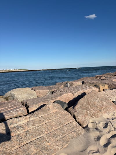 The image shows a rocky shoreline with large granite boulders in the foreground. The ocean is calm and blue, with a distant landmass visible on the horizon. The sky is clear and bright blue, with a single white cloud. The scene appears to be a public beach or park area. There is a shadow of a person on the left side of the frame, suggesting a visitor to the location. The environment is natural and serene, with a sense of open space and a clear view of the water and sky. The location is identified as Hampton Bays, United States of America.