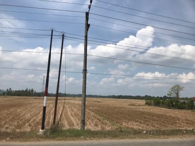 The image depicts a rural scene in Keudebagok Dua, Indonesia. A harvested rice field dominates the foreground, with stubble covering the ground. Three utility poles stand in the frame; two are dark, and one is gray. Overhead power lines crisscross the sky, partially obscuring a blue sky with scattered white clouds. In the distance, a line of trees marks the horizon, and a small red and white structure can be seen. A road borders the bottom of the frame.