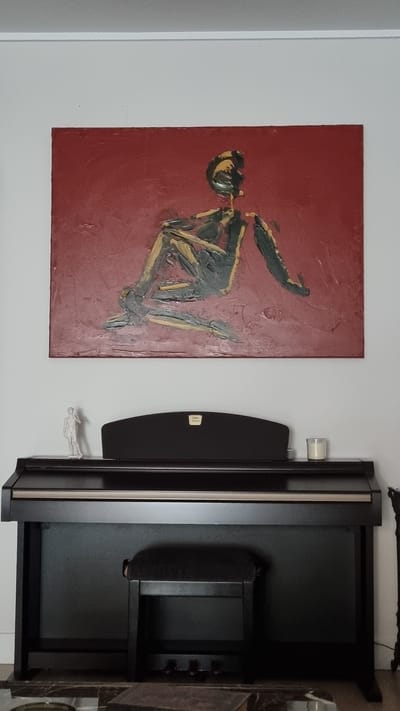 The image depicts an indoor scene featuring a dark brown digital piano with a black bench. Above the piano, a large, textured painting with a red background and an abstract figure in dark gray and yellow is displayed on a white wall. To the left of the painting, a small white figurine of a man is visible on top of the piano. A white candle in a clear glass holder sits on the right side of the piano's surface. The lighting is soft and appears to come from above. The image does not contain any specific visual cues or landmarks that would allow for the identification of its location within Jerez, Spain.
