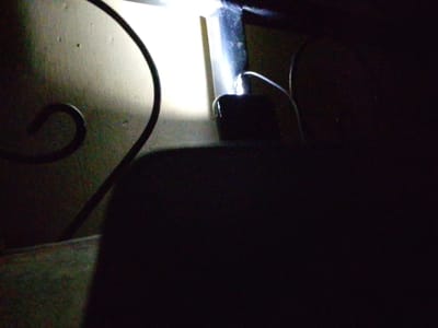 A dim room features an ornate wrought iron design on the left. A bright, white light shines from behind a dark object, possibly a piece of furniture, in the background. An indistinct form occupies the foreground, obscuring much of the scene. A wire is visible, connecting to a small, dark box, possibly an electronic device. The overall lighting is poor, making it challenging to discern specific details.