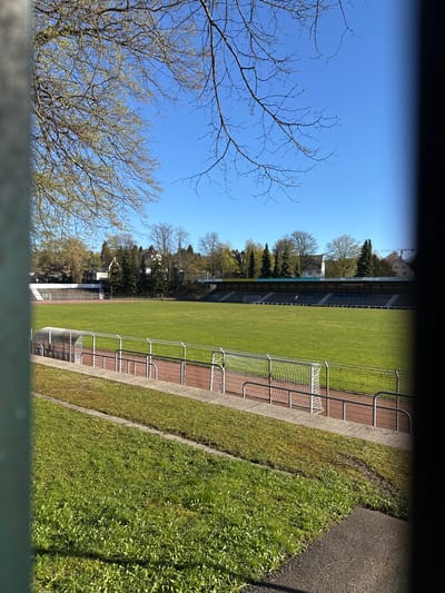 A daytime, medium-shot image from an elevated position looking towards a soccer field in Remscheid, Germany. The soccer field is green, with a running track alongside it. A metal railing separates the track from the field, and a goal is visible on one end. The stadium seating is visible on the opposite side of the field, with trees and buildings behind it. A partly bare tree is in the foreground on the left side of the frame. The sky is blue. The image is taken through a fence or barrier.