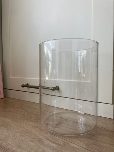 A clear, cylindrical glass container rests on a light-colored wooden floor, positioned horizontally. The container appears to be made of thick glass and has a domed end on the right side. Two metal handles, possibly made of brass or antique silver, are attached to the outside of the glass. The handles have a decorative, knob-like end and are connected to the glass by screws or rivets.
Behind the glass container is a white surface, likely cabinet doors, with visible paneling lines and a subtle sheen that reflects the light. The overall scene is indoors, suggested by the presence of the cabinet and the floor. The lighting appears to be bright, casting reflections on the glass and highlighting the texture of the wooden floor. The image is taken from a slightly elevated angle, looking down at the object. There are no people visible in the image. The environment suggests a domestic setting, possibly a kitchen or living area. There is no visible text in the image.