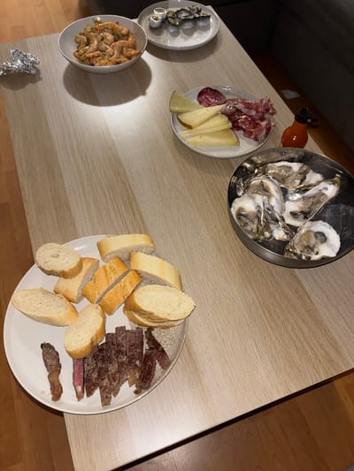 A dinner party is underway at Finca Garcés, Spain, with a spread of seafood, meats, and bread laid out on a light wood table. In the foreground, a plate showcases sliced steak and pieces of baguette, while a separate dish holds a collection of oysters on ice. To the left, a bowl is filled with cooked shrimp, and a crumpled piece of aluminum foil rests nearby. Further back, a plate displays cheese and cured meats, and another holds what appears to be sushi. A small orange bottle, possibly of hot sauce, is also visible. The scene is illuminated by ambient lighting, casting shadows on the table.