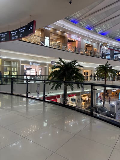 Captured from an elevated perspective, the image depicts the interior of a multi-story shopping mall. The mall features multiple floors lined with retail stores, each distinguished by unique branding and merchandise displays. Signage, including directional signs for lobbies, is visible. Palm trees are placed within the mall's central area. The mall has white tile flooring with reflective surfaces. Storefronts include recognizable brands like "UBS Gold," "Cindy," "myfeet," "erafone," and "Restowalk." The mall is located in Kelapa Dua, Indonesia.
