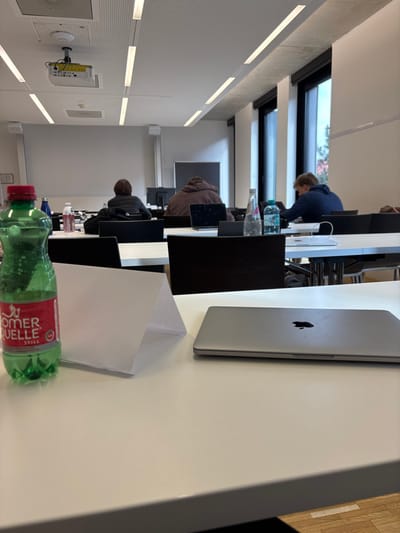 The image displays the interior of a modern classroom or lecture hall situated in Krems an der Donau, Austria. In the foreground, a white rectangular table holds a partially full green plastic bottle of Römerquelle Still water with a red cap and label on the left. Adjacent to it is a folded white piece of paper. To the right, a closed silver Apple MacBook laptop with a visible dark Apple logo rests on the table.
In the midground, three individuals are seated at white tables with dark chairs, viewed from behind. The leftmost person wears a dark jacket, facing a dark monitor. The central person wears a brown hooded sweatshirt, positioned before an open laptop and a dark monitor, with a clear glass bottle and a blue plastic bottle on their table. The rightmost person wears a blue hooded sweatshirt, situated near tall windows, and is looking downwards.
The background features a white acoustic panel ceiling with multiple linear LED light fixtures and a ceiling-mounted projector in the upper left quadrant. A large white projection screen is visible on the front wall. The right wall contains tall, dark-framed windows that admit natural light, with faint green foliage visible outside. The individuals appear independently engaged in quiet work or study; no direct interactions are observed.